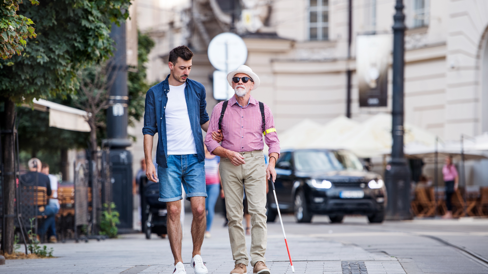 two men walking on the sidewalk. one of the men is walking with a white cane