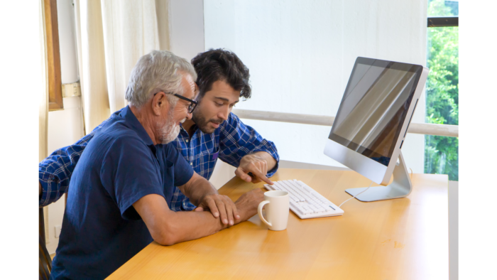 A younger man and older man sit in front of a computer. The younger man is pointing to the computer keyboard, showing the older man sitting next to him, how it works. 