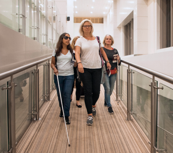 Four women walk down a hallway. The woman on the right is wearing dark shades and is using a white cane.