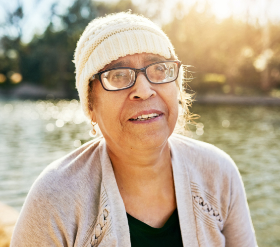 A woman wearing a white knit hat and glasses smiles while the sun shines behind her.