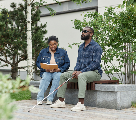 A woman reads to a man wearing dark shades and holding a white cane while both are seated in a park.