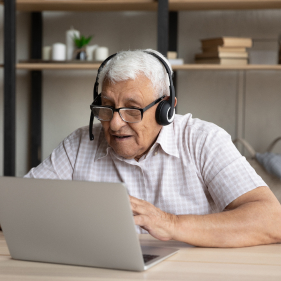 Elderly man with white hair wearing glasses and headphones watching the screen of a laptop.