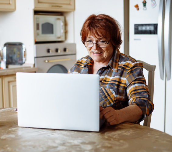 A woman with red hair and glasses uses a laptop while seated in her kitchen.