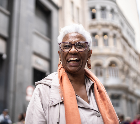 Woman with short white hair, glasses and peach colored scarf smiling outside with city buildings in the background.