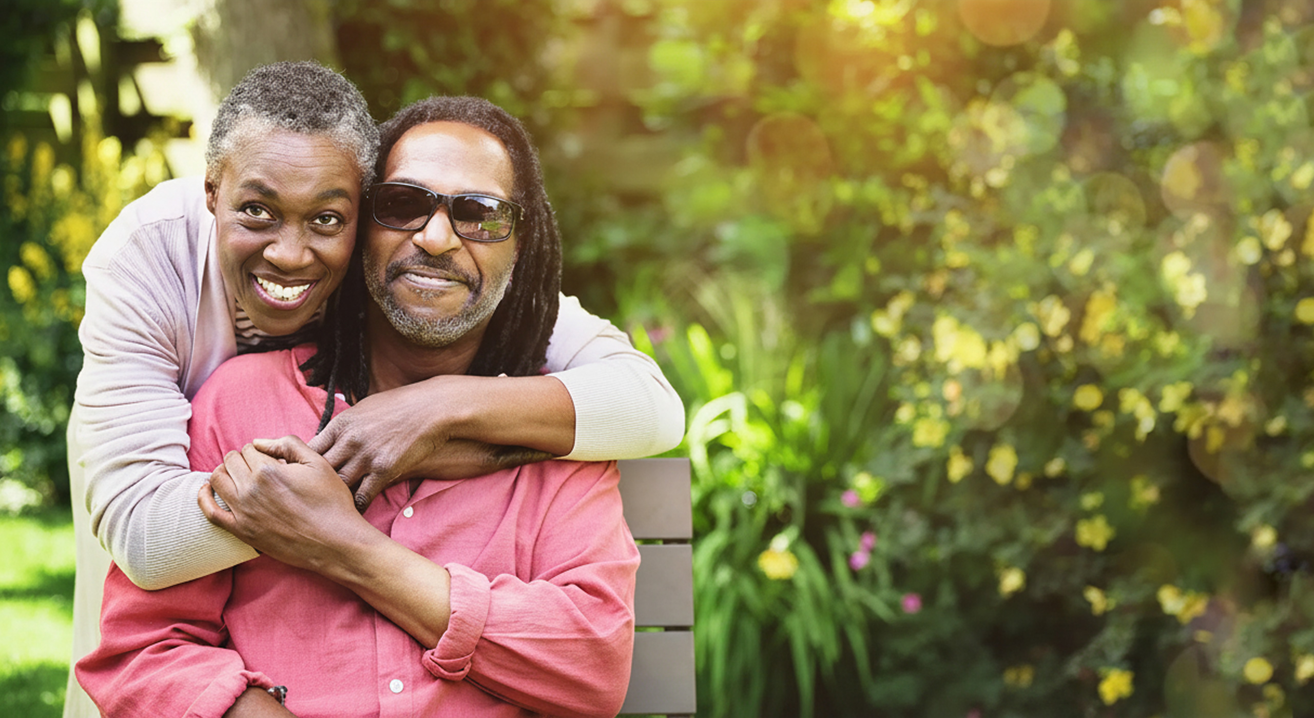 A woman with her arms wrapped around a man wearing dark sunglasses, sitting in a garden.