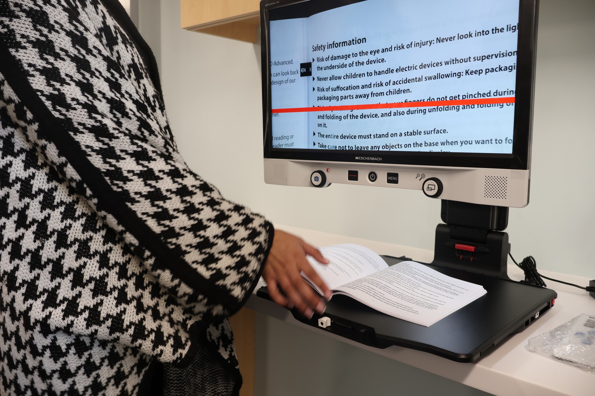 A person uses a desktop video magnifier to read printed text from a book, with the magnified text displayed on the monitor.