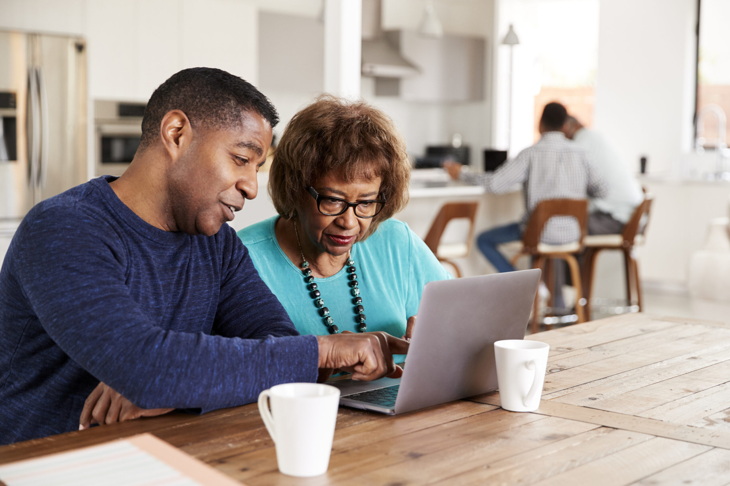 A man and woman sit at a table in a bright and modern room, focused on a laptop. The man points to the screen, assisting the woman.