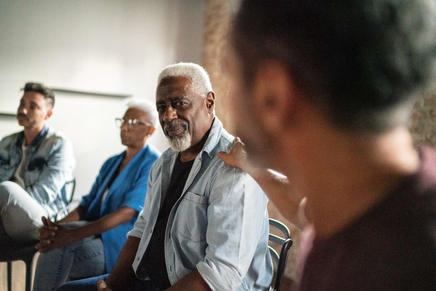 A diverse group of people sit in a circle during a support meeting. An elder man with a gentle smile is comforted by a hand on his shoulder.