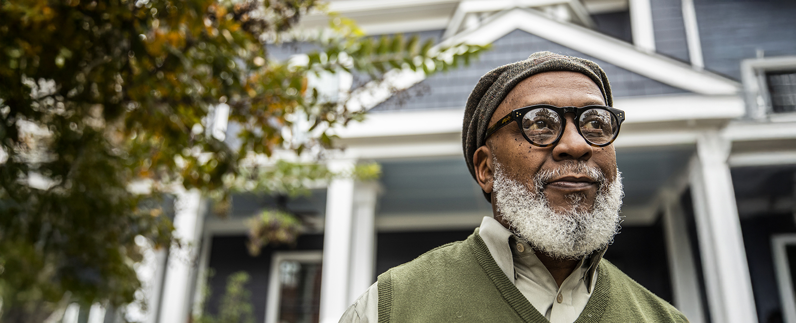 A man with glasses stands confidently outside a blue house. He appears thoughtful and content.