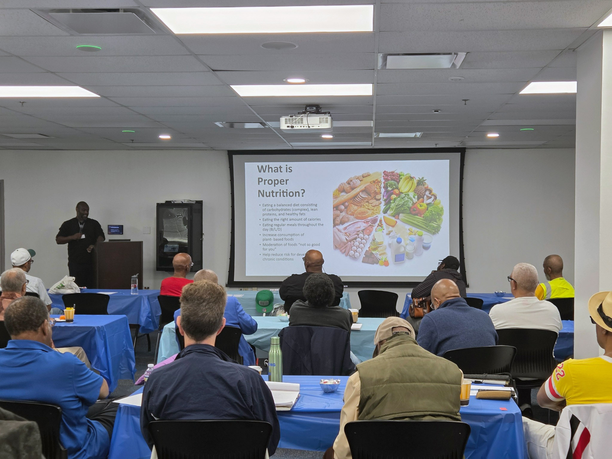 A speaker presents a nutrition slide to a diverse group of a dozen men in a conference room.