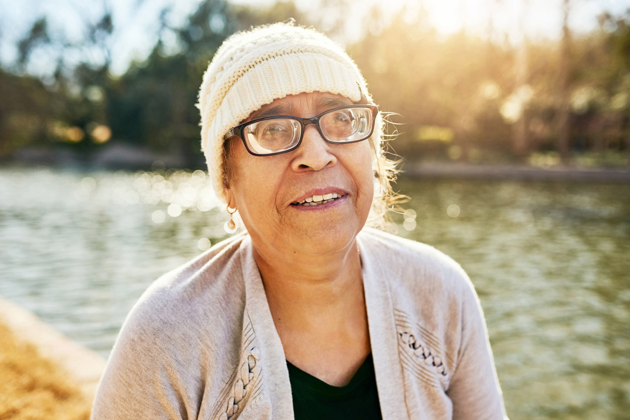 A woman wearing a white knit hat and glasses smiles while the sun shines behind her.