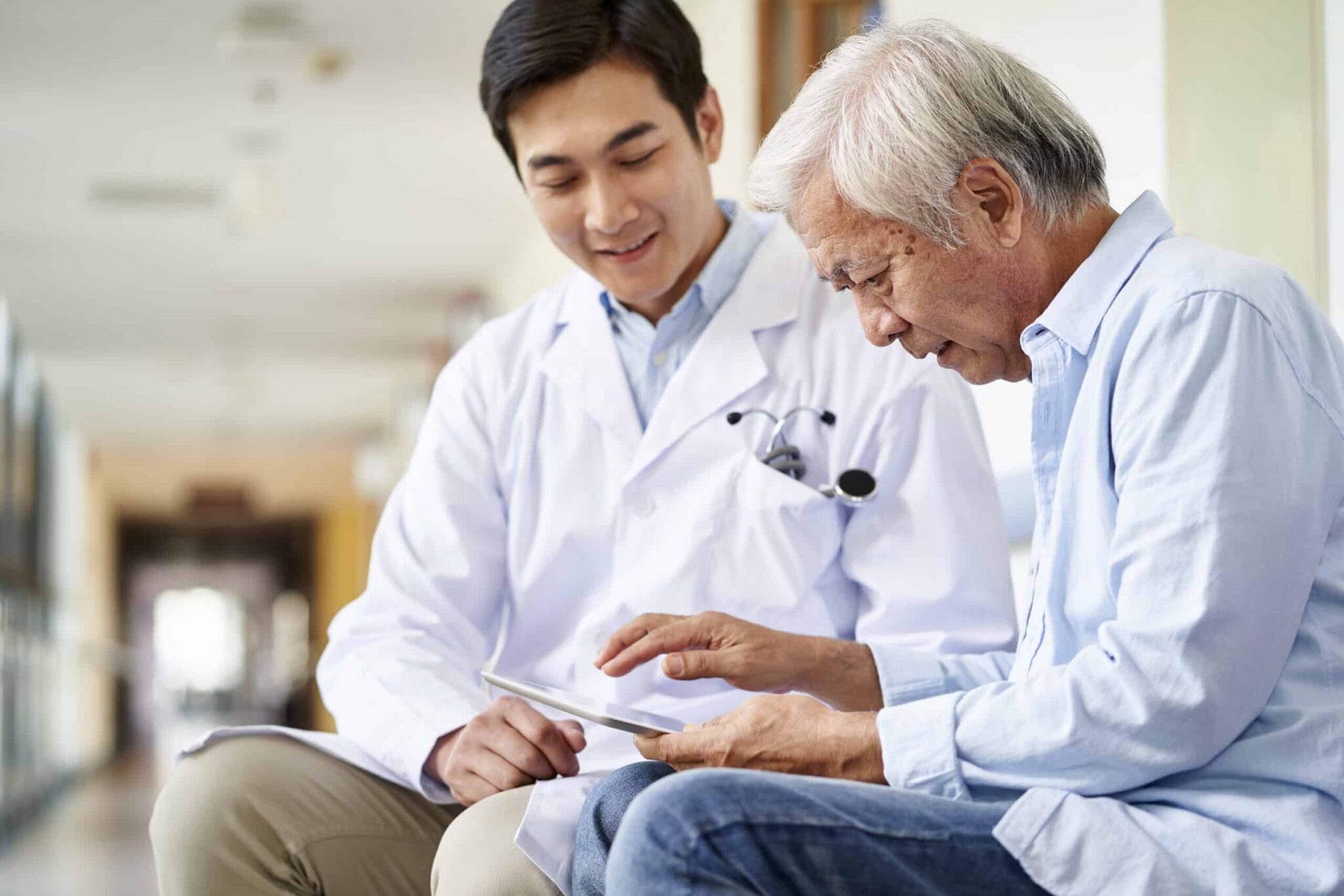A doctor in a white coat sits beside an elderly man, who is using a tablet.