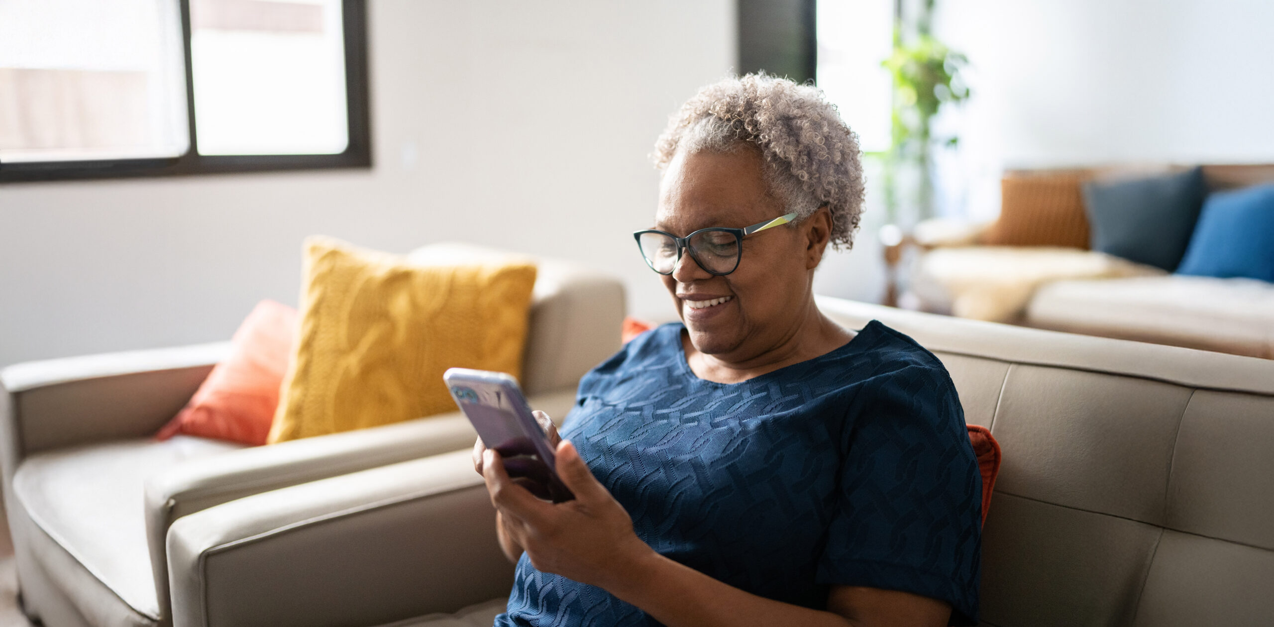 A woman sitting on a beige sofa in a bright, cozy room, wearing glasses and smiling while using a smartphone.
