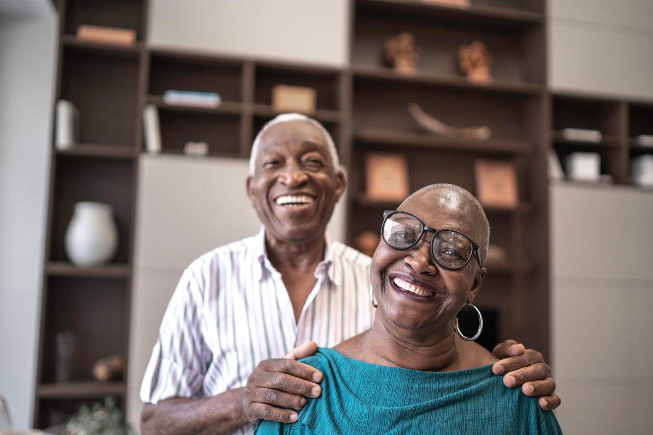 Two seniors in a casual embrace, wearing glasses and smiling at the camera.