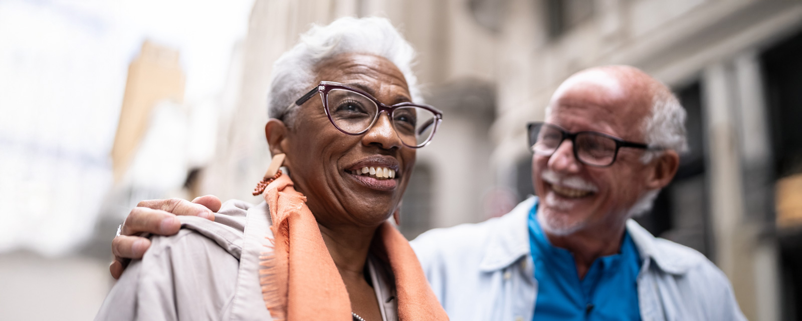 Two friends wearing glasses smiling warmly while walking in a city together.