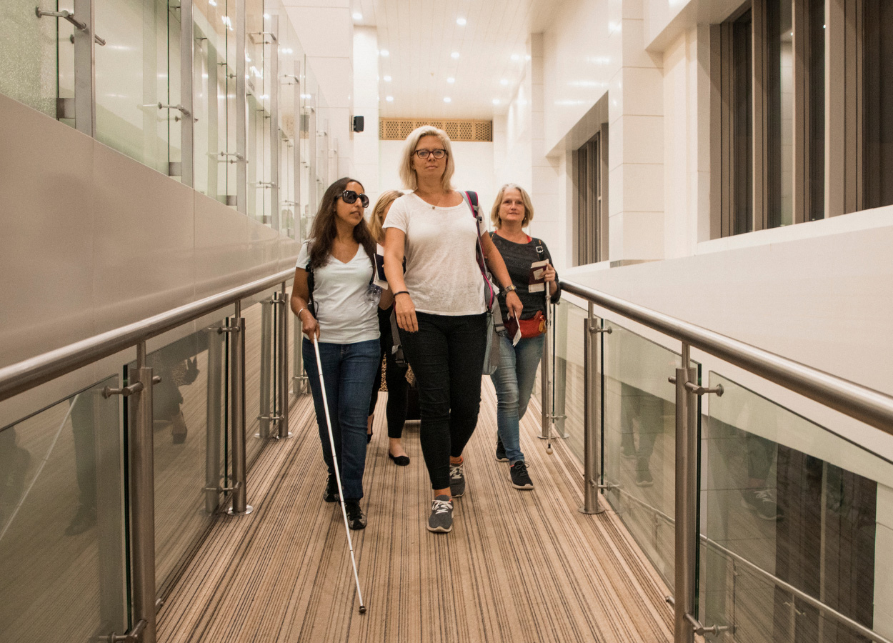A woman using a white cane walks confidently down a well-lit corridor, flanked by supportive companions.
