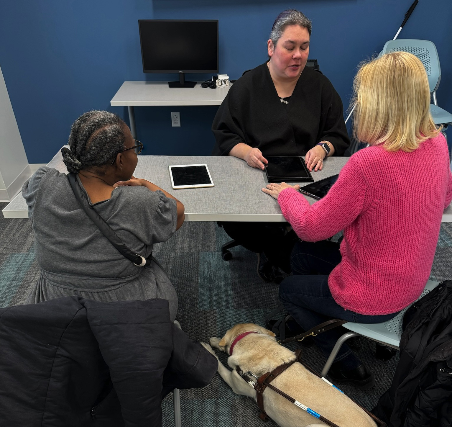 An access technology instructor and two people sit at a table with tablets, engaged in conversation. A guide dog rests on the floor.