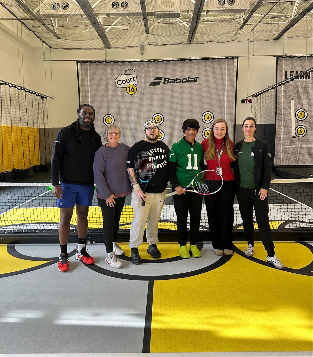 Six people pose in front of a tennis net on an indoor court at Court 16. Two of the people are holding tennis rackets.