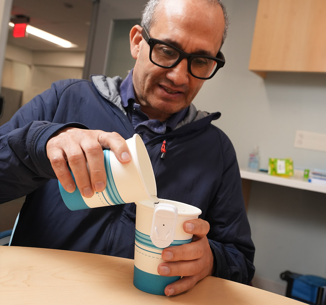 A man in a navy jacket seated a table using a liquid leveler during a demonstration at VisionLink, pouring liquid from one cup into another cup that has a small white device attached to its rim.