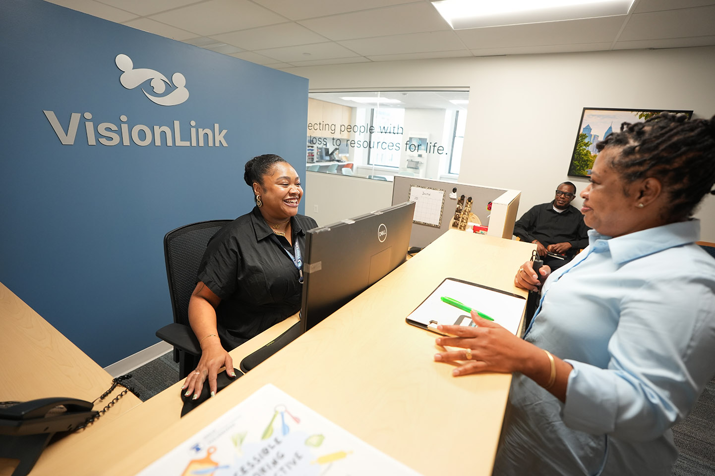 A receptionist is seated at her desk with a blue wall behind her, smiling and welcoming a VisionLink client.