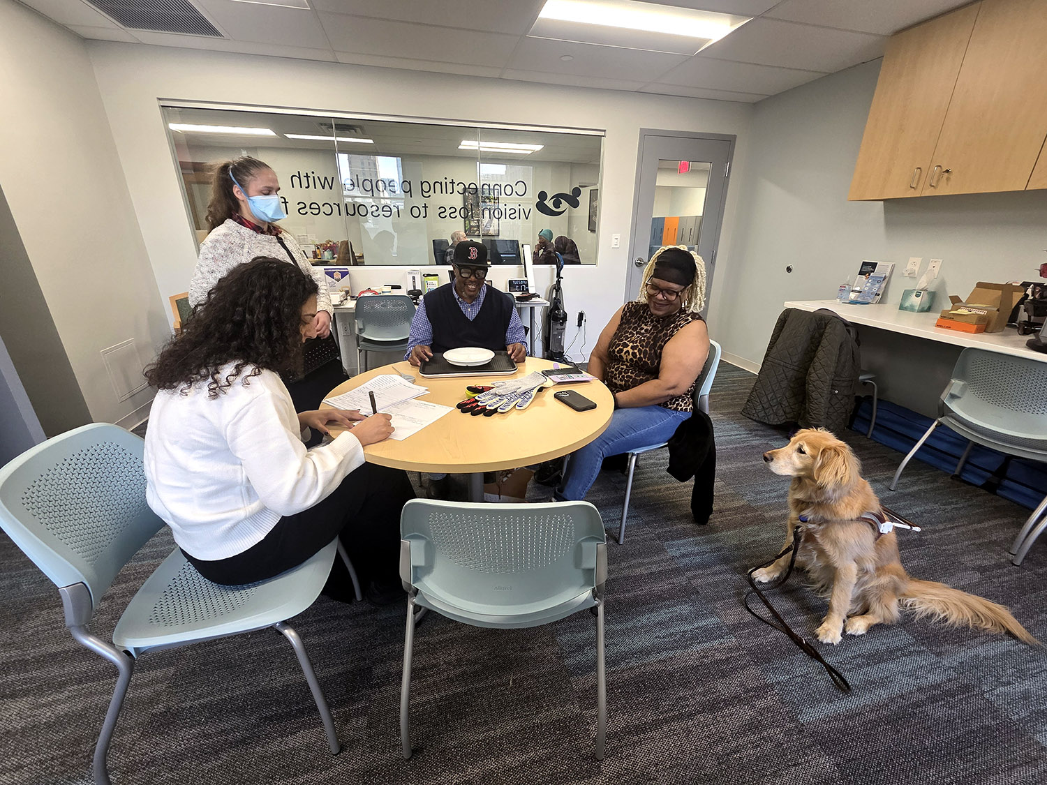 A group of people are seated around the table in VisionLink's Low Vision Resource Center. On the table are assistive cooking tools and paperwork. To the right, a guide dog sits at attention.