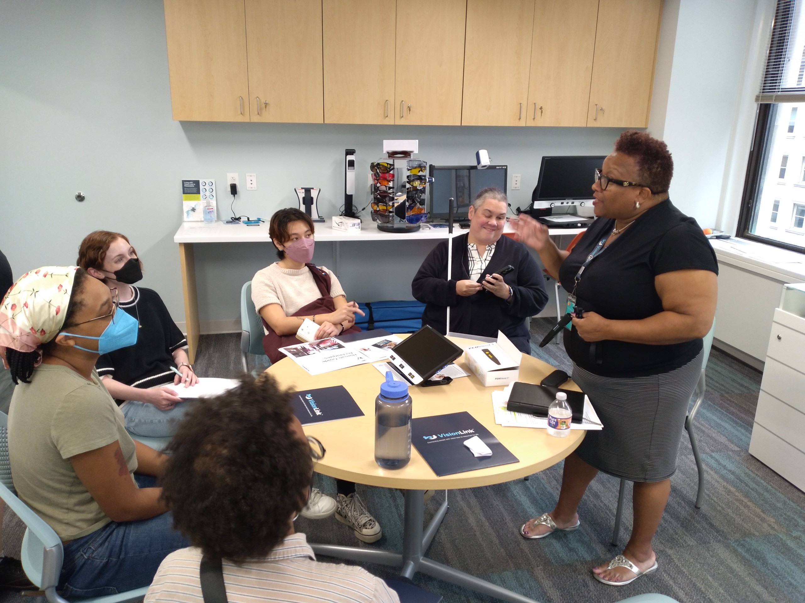 Members of Art Reach are seated at a round table in VisionLink's Low Vision Resource Center while a staff member stands and presents to them.