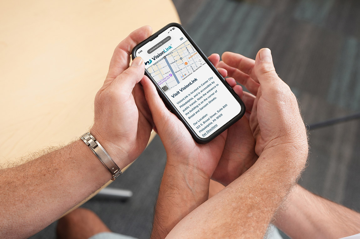An instructor guiding a student's hands on a smartphone displaying a VisionLink web page with a map and location details for their Philadelphia office.