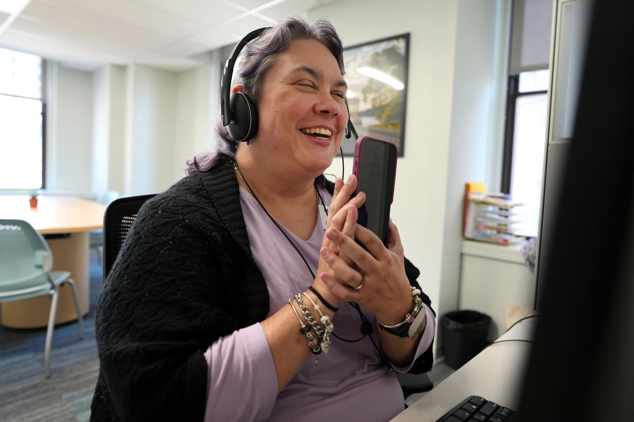 A woman, wearing a headset and turning her iPhone screen to the camera, smiles and laughs during a video call at a desk in a VisionLink meeting room; the mood is warm and joyful.