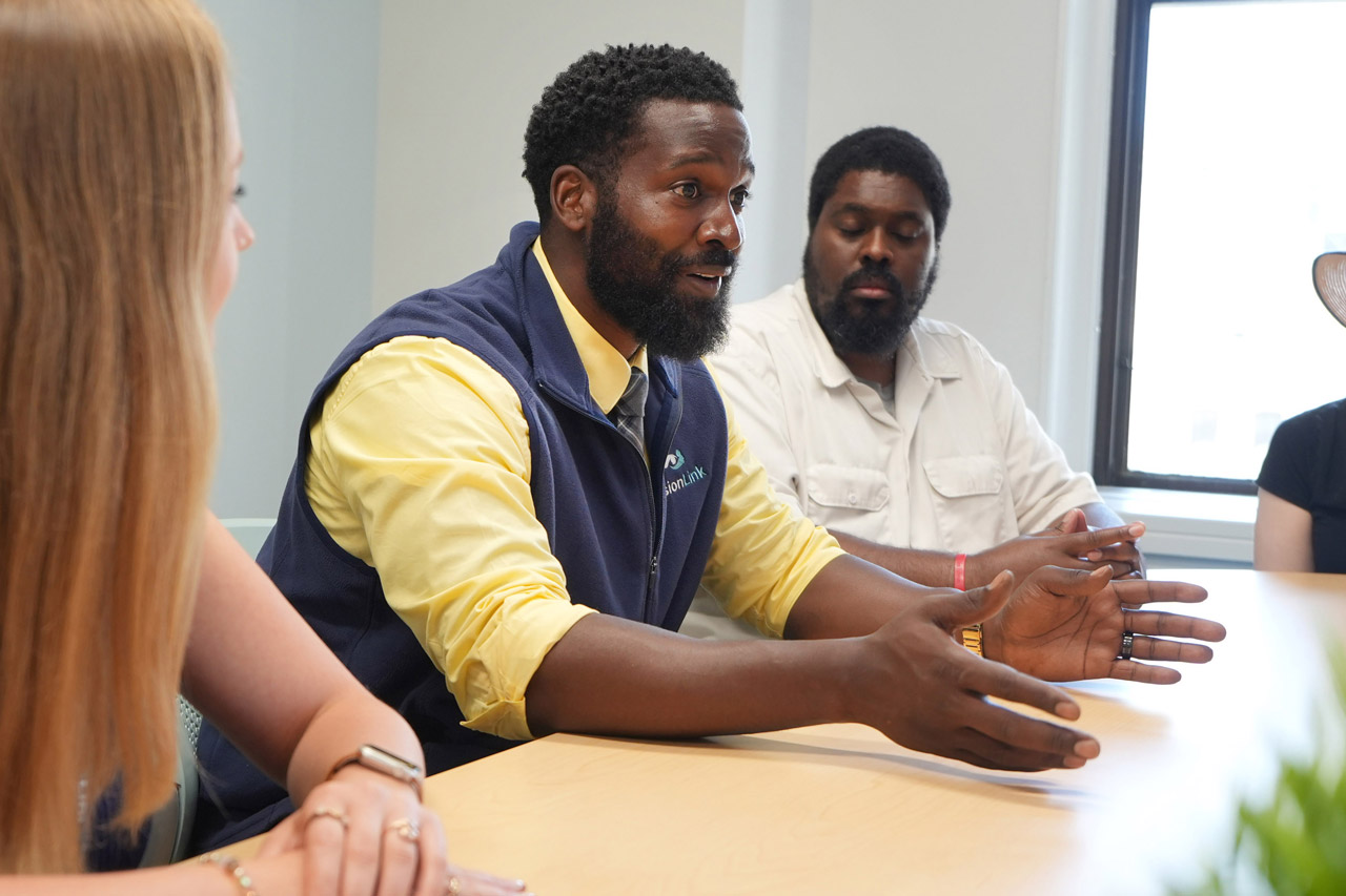 A VisionLink staff member in a yellow shirt and navy vest speaks with animated hand gestures at a classroom table as colleagues listen, creating an engaged, collaborative tone.